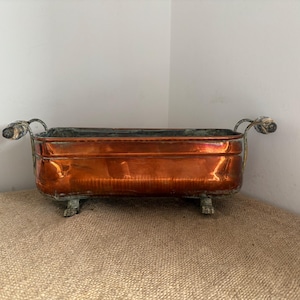 Puede incluir: Una jardinera rectangular de cobre con acabado pulido, que presenta dos asas metálicas curvas en los laterales y cuatro patas pequeñas en la base. El interior muestra signos de oxidación y el exterior exhibe un tono naranja metálico cálido.
