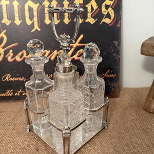 May include: A vintage perfume bottle set with three clear glass bottles with crystal stoppers, set in a silver-toned metal tray with a handle. The bottles have intricate cut glass designs. The background includes a vintage sign.