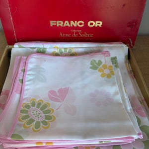 May include: A vintage bedding set featuring white sheets and pillowcases with a floral pattern in pink, green, and yellow. The set is neatly folded inside a red box with the text "FRANC OR Collection Anne de Solène".