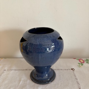 May include: A blue ceramic vase with a unique design featuring cutouts on the sides. The vase has a rounded body and a flared base.