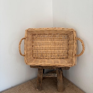 May include: A rectangular woven wicker basket with a light tan natural finish. The basket features a sturdy base, vertical side walls, and two rounded handles attached to the sides. It is positioned on a small, dark brown wooden stool.