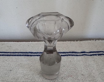 May include: A clear glass bottle stopper with a faceted, rounded top and a cylindrical base. The stopper is transparent, allowing light to pass through, and sits on a white and blue striped fabric.