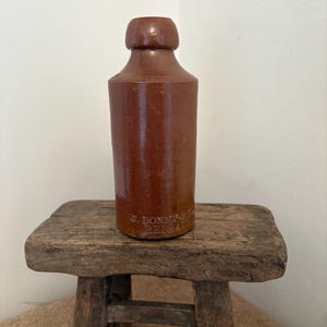 May include: A brown stoneware bottle with a rounded top and cylindrical body. The bottle has a dark brown color and the text "J. BONN & S. REIGATE" is printed on the front. The bottle is displayed on a small, rustic wooden stool.