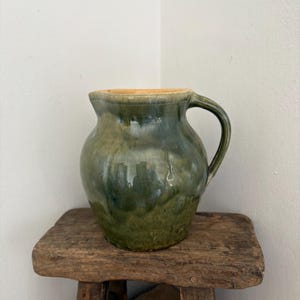 May include: A green glazed ceramic jug with a handle. The jug has a rounded body and a slightly flared rim. It sits on a small, rustic wooden stool. The jug's glaze has variations of green hues.