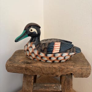 Puede incluir: Una figura de pato de madera pintada a mano, con cabeza negra, pico turquesa y detalles de plumas en tonos marrón, naranja y azul. El pato descansa sobre un taburete de madera rústico y desgastado, mostrando texturas y colores naturales.