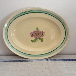 Könnte beinhalten: Ovale Servierplatte mit cremefarbener Oberfläche, mit einem Blumendesign in der Mitte. Die Platte hat grüne Streifen um den Rand. Das Blumendesign umfasst rosa und lila Blumen mit grünen Blättern.