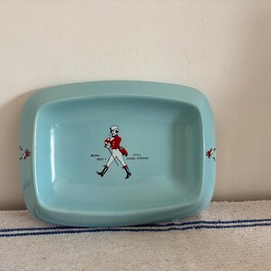 May include: A light blue rectangular ceramic dish with rounded corners. The dish features a graphic of a man in a red coat and white pants, with the text "BORN 1820 - STILL GOING STRONG". Decorative red accents are on the sides.