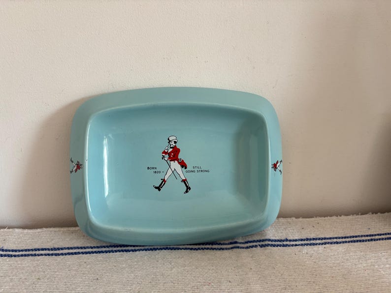May include: A light blue rectangular ceramic dish with rounded corners. The dish features a graphic of a man in a red coat and white pants, with the text "BORN 1820 - STILL GOING STRONG". Decorative red accents are on the sides.