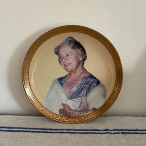 May include: A decorative plate with a portrait of a woman with grey hair, wearing a blue dress and jewellery. The plate has a gold rim with an ornate design and the years "1900 2002" are printed on the plate.