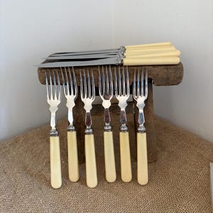 May include: A collection of vintage silverware, including forks and knives, with cream-colored handles and silver blades. The forks are arranged in a row, while the knives rest on a wooden surface. The set is displayed on a rustic wooden stool.