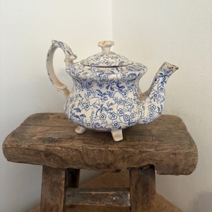May include: A vintage teapot with a blue floral pattern on a white background. The teapot has a curved handle, a spout, and a lid with a small knob. It is resting on a rustic wooden stool. The teapot is a charming piece of kitchenware.