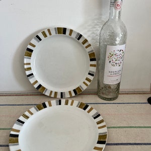 Peut inclure: Deux assiettes en c&eacute;ramique blanche avec des bordures ray&eacute;es noires, dor&eacute;es et grises. Les assiettes sont sur une nappe ray&eacute;e. Une bouteille en verre transparent avec une &eacute;tiquette indiquant "Sou&ccedil;on de Fruit" est &agrave; l'arri&egrave;re-plan.