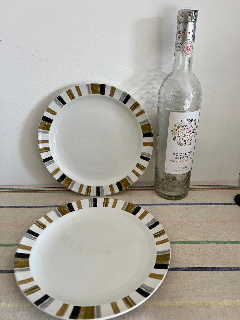 Peut inclure: Deux assiettes en c&eacute;ramique blanche avec des bordures ray&eacute;es noires, dor&eacute;es et grises. Les assiettes sont sur une nappe ray&eacute;e. Une bouteille en verre transparent avec une &eacute;tiquette indiquant "Sou&ccedil;on de Fruit" est &agrave; l'arri&egrave;re-plan.