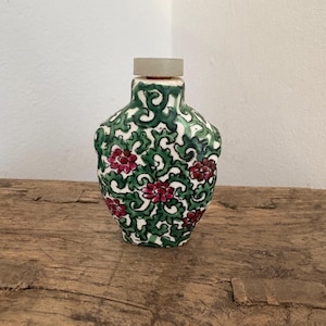Puede incluir: Pequeña botella decorativa de cerámica con diseño floral. La botella presenta un fondo verde y blanco con flores rojas. Tiene un tapón claro y está sobre una superficie de madera.