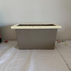 Pode incluir: Um vaso retangular de metal cinza com uma borda branca. O vaso está sobre uma toalha de mesa branca.