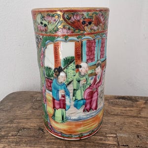 May include: A cylindrical porcelain vase featuring a hand-painted scene of three figures in traditional attire within an architectural setting. The design includes vibrant colors such as pink, green, blue, and red, with gold accents along the rim.