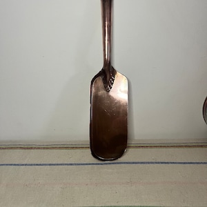 May include: A copper fireplace shovel with a long handle. The shovel has a decorative design at the top of the handle.