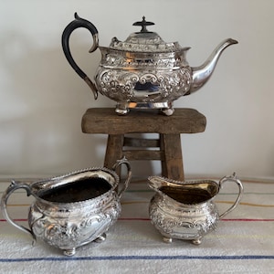 May include: A silver tea set with ornate floral detailing. The set includes a teapot with a black handle, and two creamers. The teapot sits on a small wooden stool. The set is displayed on a striped tablecloth.