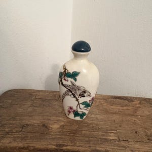 Puede incluir: Una botella de cerámica color crema con un tapón azul oscuro. La botella está decorada con una ilustración detallada de un pájaro posado en una rama floreciente con flores rosas y hojas verdes. La botella se exhibe sobre una superficie de madera rústica.