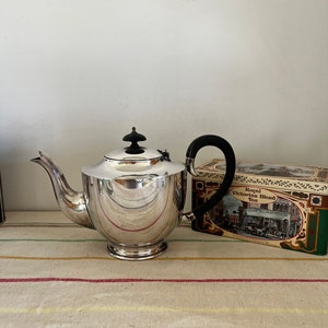 Puede incluir: Una tetera de plata con asa negra y un pomo negro en la tapa. La tetera está sobre un mantel a rayas junto a una caja de té Royal Victorian Blend.