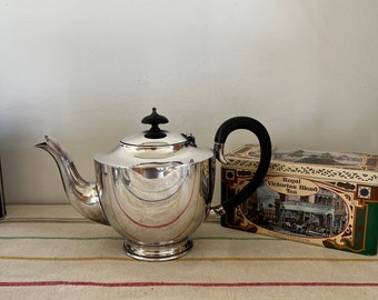 May include: A silver teapot with a black handle and a black knob on the lid. The teapot is sitting on a striped tablecloth next to a box of Royal Victorian Blend Tea.