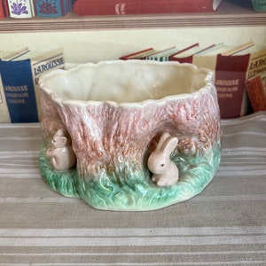 Könnte beinhalten: Ein Keramik-Pflanztopf in Form eines Baumstamms mit zwei kleinen Hasen an der Seite. Der Topf ist mit brauner, grüner und rosa Glasur verziert.