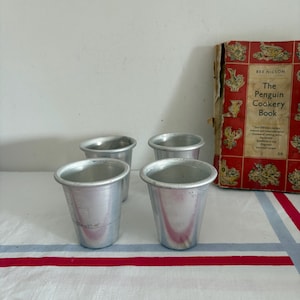 May include: Four small, silver metal cups with a slightly tapered design are arranged on a white tablecloth with red and blue stripes. A vintage cookbook titled "The Penguin Cookery Book" is visible in the background.
