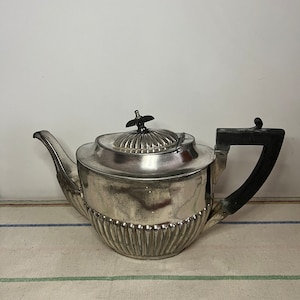 Puede incluir: Una tetera de plata con asa negra y diseño acanalado. La tetera tiene un cuerpo redondo y una tapa abovedada con un pomo en la parte superior.