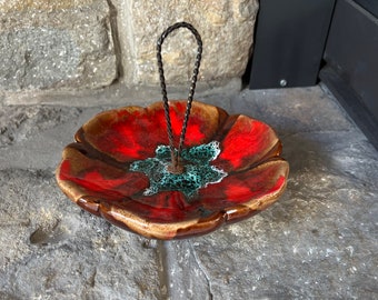 May include: A red and brown ceramic flower-shaped dish with a green center and a black metal handle. The dish is sitting on a stone surface.