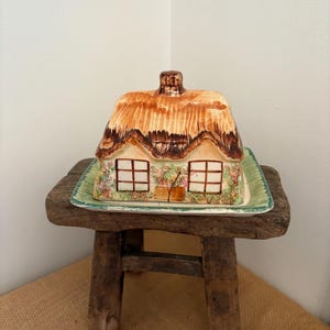 May include: A ceramic cottage-shaped butter dish with a thatched roof and two windows. The dish sits on a rectangular base with a green border. The cottage is brown, beige, and white. The dish is on a small, rustic wooden stool.