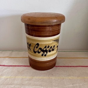 Może przedstawiać: Ceramiczny pojemnik na kawę w kolorze brązowym i beżowym z drewnianym wieczkiem. Pojemnik ma brązowo-beżowe pasy z czarnym napisem "Coffee" i wzorem liści.