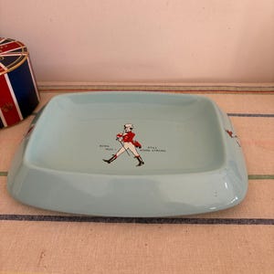 May include: A light blue ceramic ashtray featuring a red and white illustration of a man in a red coat and black boots walking with a cane. The text "BORN 1820" and "STILL GOING STRONG" is printed on the ashtray.