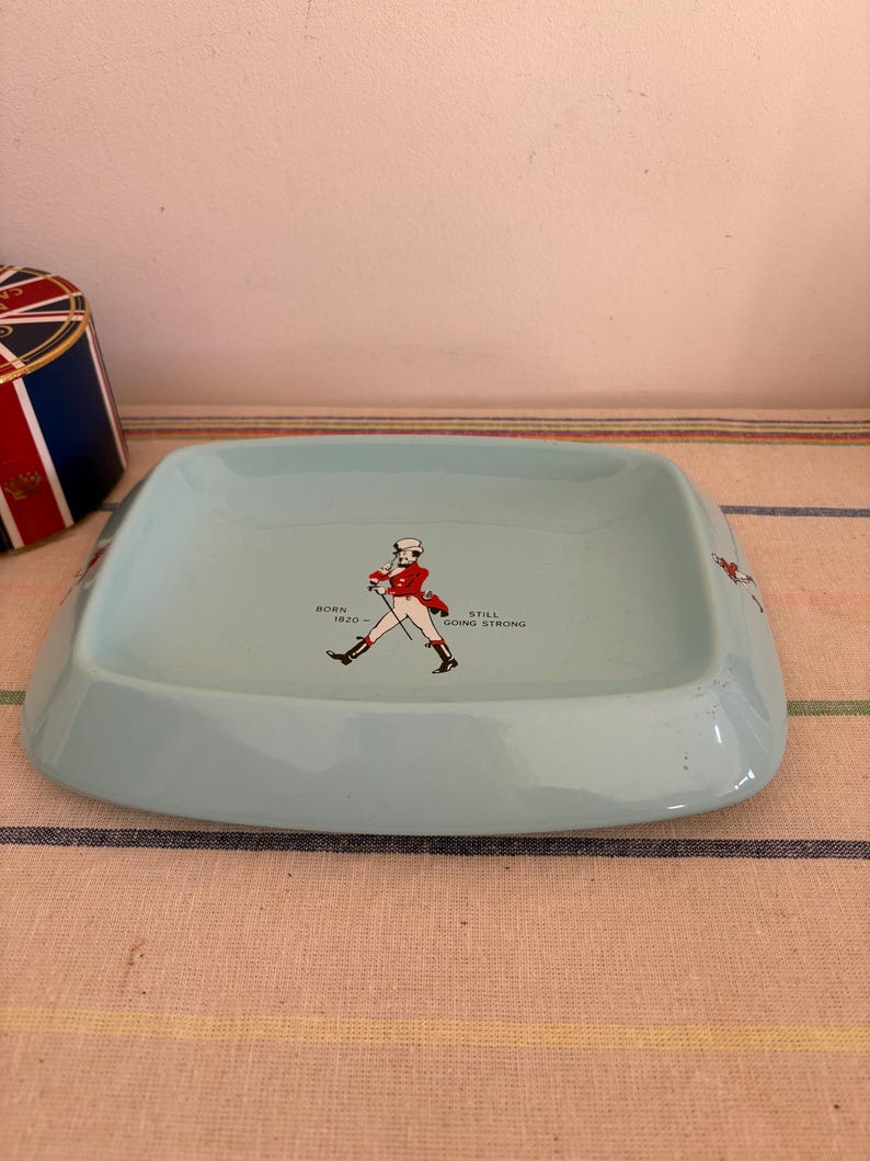 May include: A light blue ceramic ashtray featuring a red and white illustration of a man in a red coat and black boots walking with a cane. The text "BORN 1820" and "STILL GOING STRONG" is printed on the ashtray.