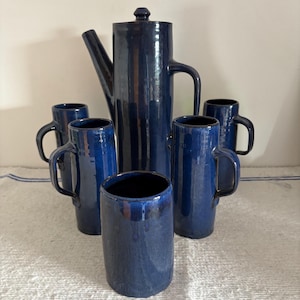 May include: A set of cobalt blue ceramic coffee service. The set includes a tall coffee pot with a lid, and six matching mugs with handles. The ceramic has a glossy finish. The set is arranged on a white surface.