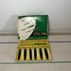 Puede incluir: Una caja de 12 tenedores de cóctel "Little Fork" de plata con mangos de plástico blanco y negro. La caja es verde y blanca y dice "Special Quality" y "Trade Mark".