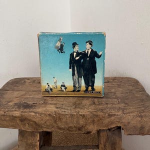 May include: Vintage box featuring a classic image of Laurel and Hardy in formal attire against a blue sky backdrop. The box has a Film Office logo and a distressed, aged appearance, suggesting it's a collectible item. The box is placed on a rustic wooden stool.