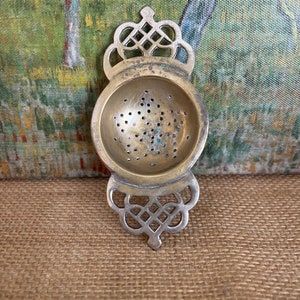 May include: An antique tea strainer with a decorative filigree design. The strainer is made of brass and features a round bowl with small holes for straining tea leaves. The handle and top are silver-colored with an ornate pattern.