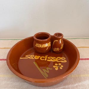 May include: A terracotta serving dish with two small terracotta pots. The dish is decorated with the words "saucisses" and "moutarde" in yellow paint. The pots are decorated with the words "moutarde" and "w.re." in yellow paint.