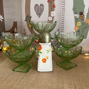 Könnte beinhalten: Satz von vier grünen Glasschalen mit geriffelten Rändern und quadratischen Böden. Die Schalen sind in einem Kreis um einen weißen Keramikstreuer mit Blumenmuster und silbernem Deckel angeordnet.
