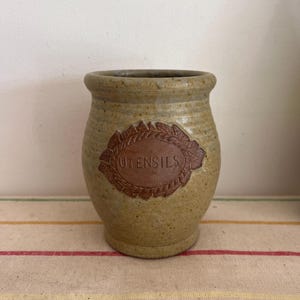 May include: A brown and tan ceramic utensil holder with a brown label that says "UTENSILS" in a white font.