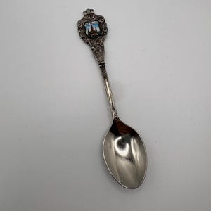 May include: A silver souvenir spoon with a detailed handle and a bowl. The handle features a colourful emblem with the text "NEW ORLEANS". The spoon is likely a collectible item.