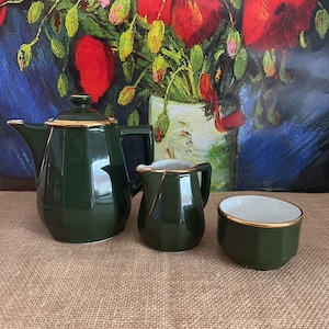 May include: A green tea set with gold trim. The set includes a teapot, creamer, and sugar bowl. The teapot has a lid and a handle. The creamer and sugar bowl have handles. The set is on a brown hessian surface.