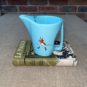 May include: A light blue ceramic creamer with a handle. The creamer features a red and white illustration of a man in a red coat walking with a cane. The text "Born 1820 - Still Going Strong" is printed on the creamer.