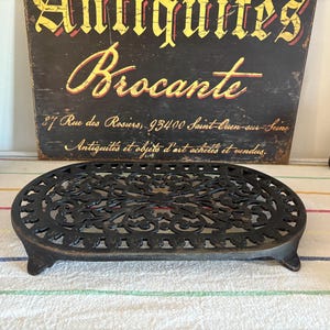Puede incluir: Salvamanteles de hierro fundido negro con forma ovalada y diseño calado ornamentado. El salvamanteles tiene pequeños pies y se exhibe sobre una tela a rayas. Un letrero vintage en el fondo dice "Antiquites Brocante".