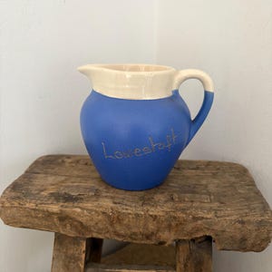 Könnte beinhalten: Blau-cremefarbener Keramikkrug mit dem Wort "Lowestoft" in goldener Schrift. Der Krug hat einen Henkel und einen Ausguss. Er steht auf einem rustikalen Holzschemel.