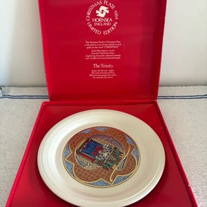 May include: A vintage Hornsea England Christmas Plate from 1984, displayed in a red presentation box. The plate has a central design with blue, red, and gold details. The box is marked "Christmas Plate" and is part of a limited edition.