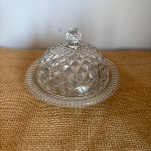 Puede incluir: Una mantequera de cristal transparente con tapa abovedada con diseño de diamantes y base redonda con borde estriado. La tapa tiene un pequeño pomo redondo en la parte superior. La mantequera está sobre una superficie marrón texturizada.