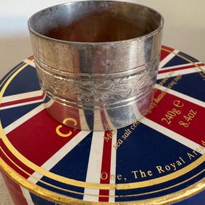 May include: Silver napkin ring with an engraved design. The ring is sitting on top of a box with a Union Jack design and the text "One, The Royal Arcade".