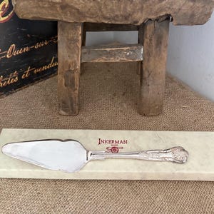 May include: A silver-plated cake server with an ornate handle featuring a shell motif, resting on a rectangular cream-colored presentation box. The box is printed with the red text INKERMAN and a small cannon icon above the word SILVER.