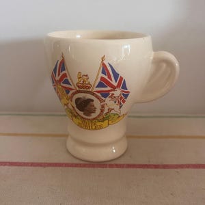 Puede incluir: Una taza de cerámica blanca con asa, con un diseño conmemorativo del Jubileo de Plata de la reina Isabel II en 1977. El diseño incluye el perfil de la reina, dos banderas británicas cruzadas y dos leones.